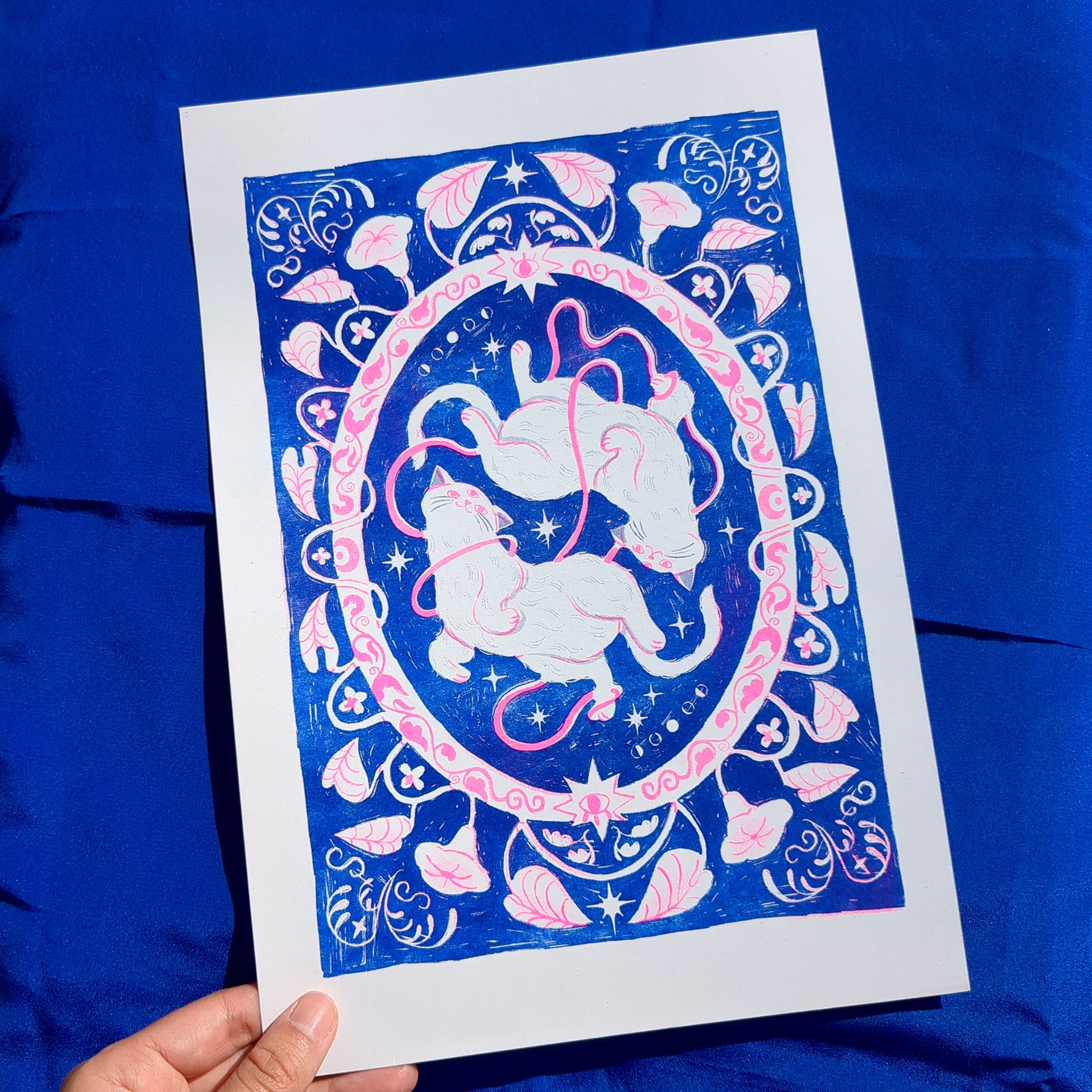 Blue and pink risograph print of a symmetrical, folk-like cat design with foliage surrounding, picked up by a hand