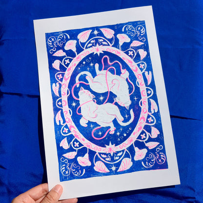 Blue and pink risograph print of a symmetrical, folk-like cat design with foliage surrounding, picked up by a hand
