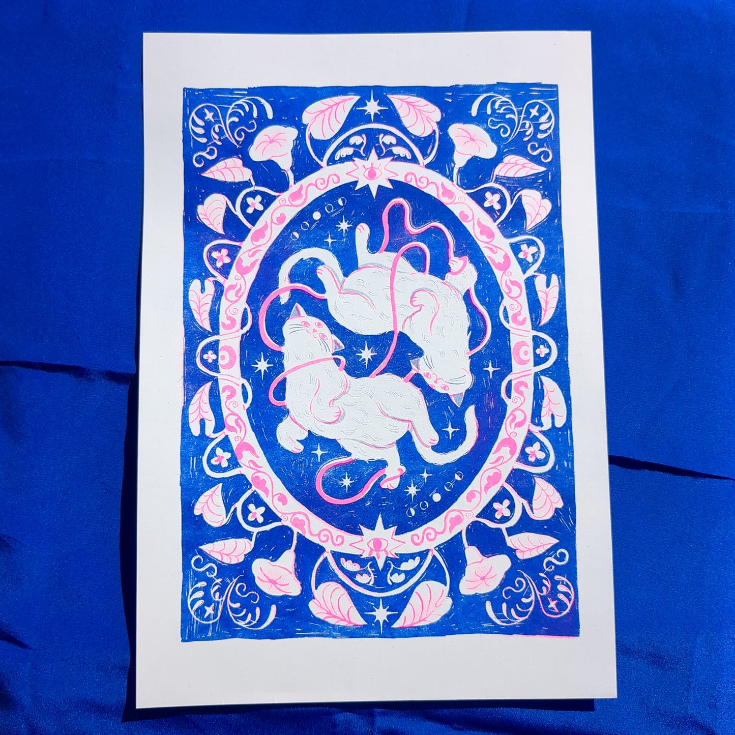 Blue and pink risograph print of a symmetrical, folk-like cat design with foliage surrounding, front view