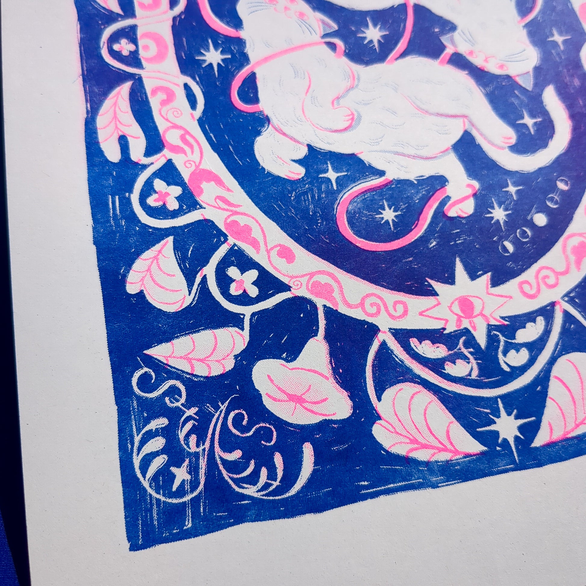 Blue and pink risograph print of a symmetrical, folk-like cat design with foliage surrounding, super close up on foliage